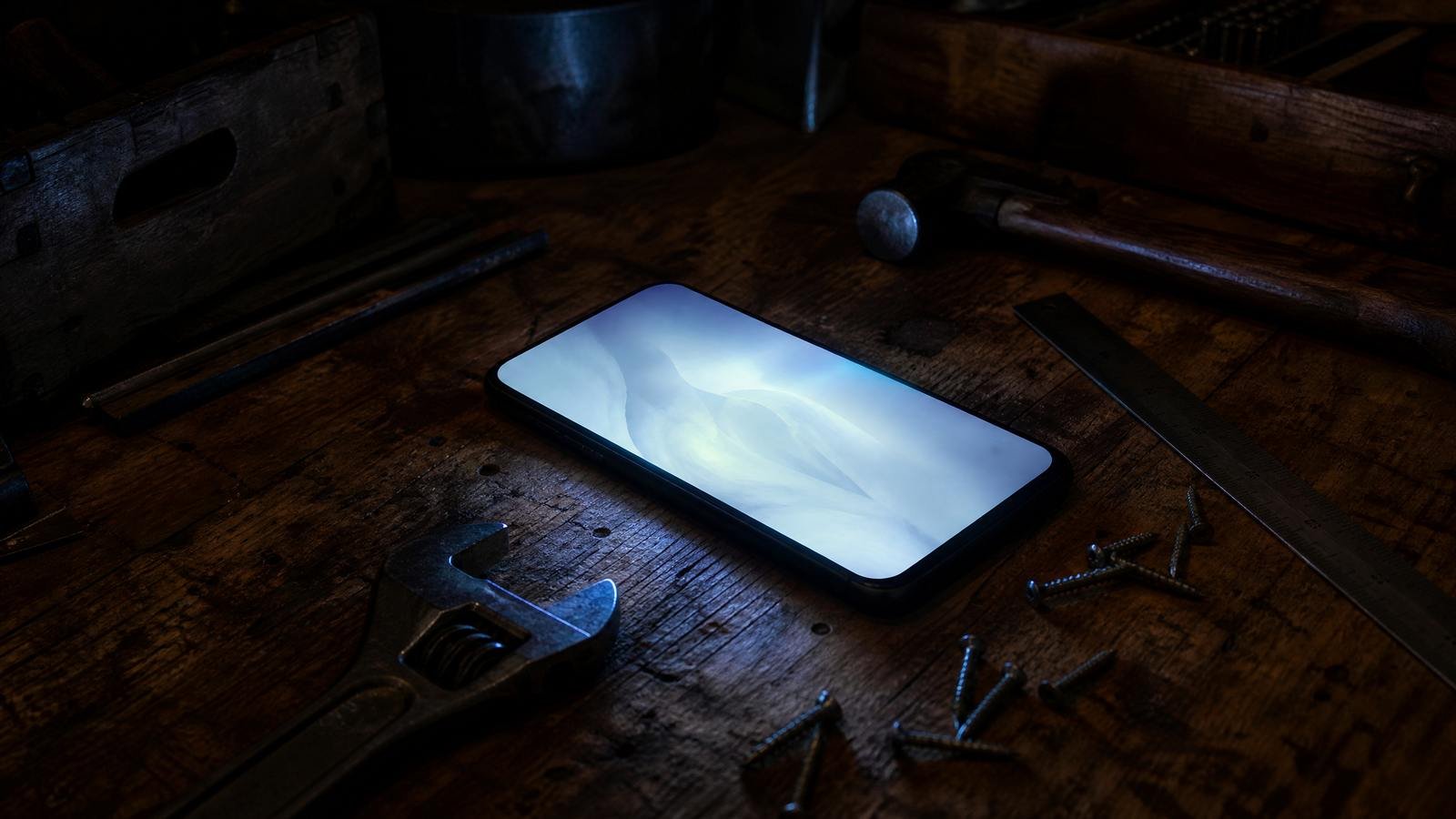 A phone lying face up and glowing on a dark workbench at night, a wrench, screws and a steel rule around it