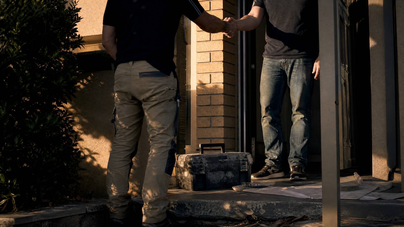 A tradesperson and a homeowner shaking hands at a suburban front door, toolbox on the step