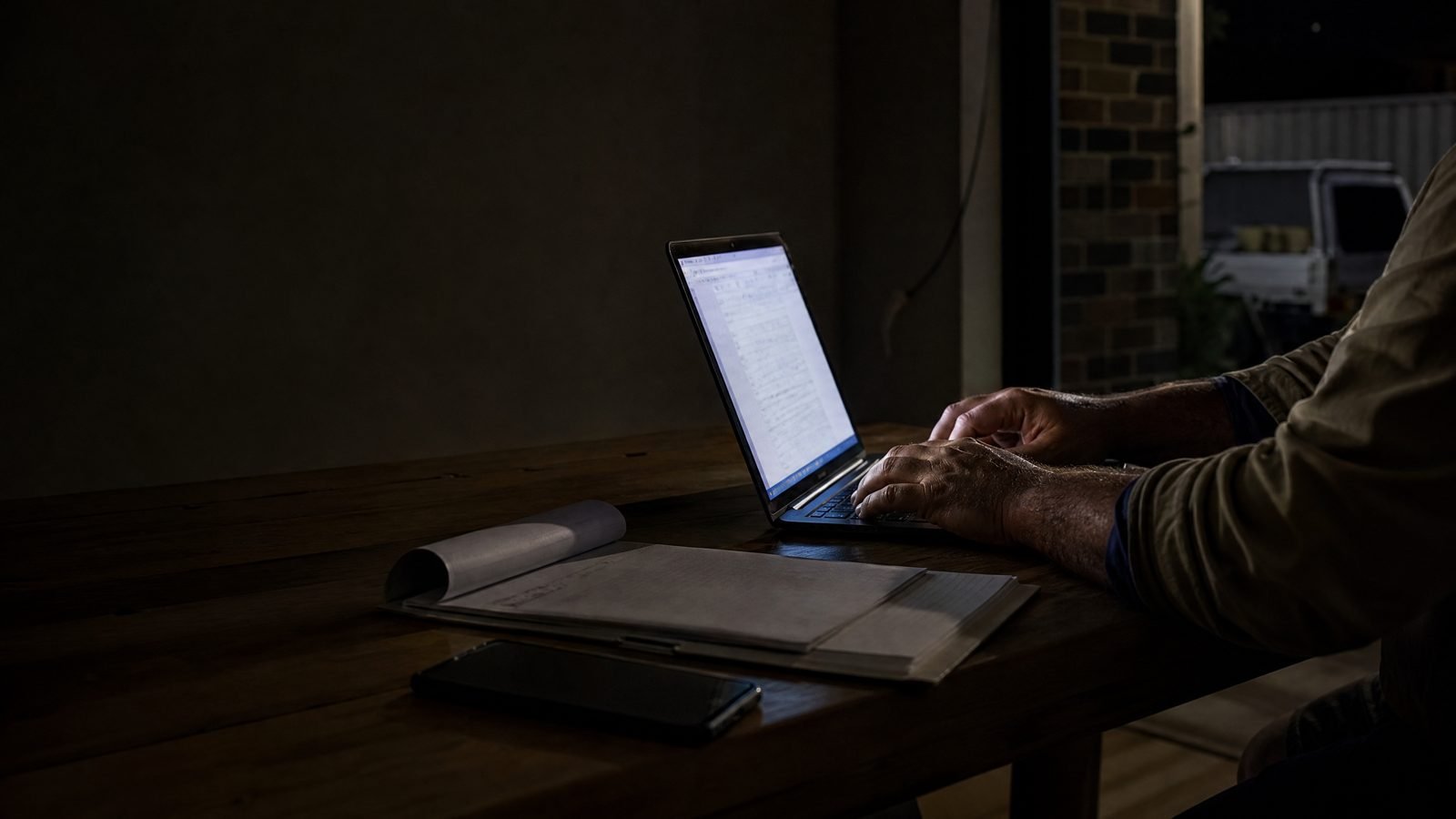 A tradesperson typing on a laptop at a timber table after dark, paperwork beside it and the ute outside