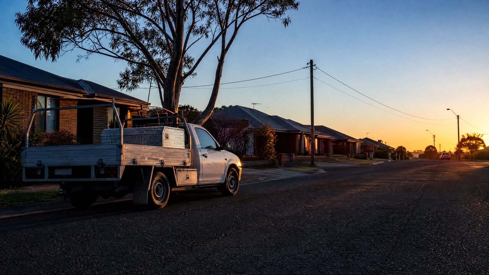A tray-back work ute parked on a quiet suburban street at sunset, gum tree overhead and the road running to an orange horizon